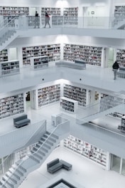 A modern library interior with multiple levels of bookshelves, staircases, and open spaces. The design is minimalist with a mostly white color scheme, allowing the colorful spines of books to stand out. The environment appears quiet and spacious, with a few people walking around.
