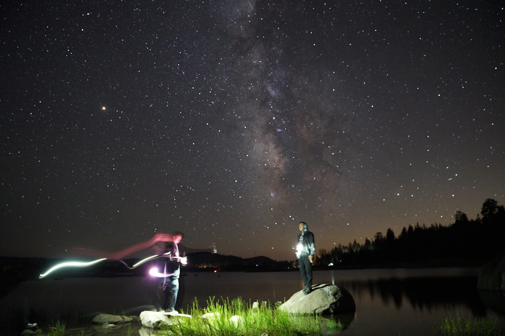 a couple of people standing next to a lake under a night sky
