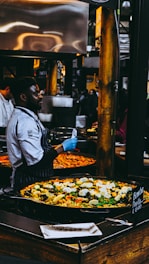 A chef wearing a striped apron and blue gloves is preparing and serving a large pan of colorful paella in a busy market setting. The paella is filled with ingredients like rice, bell peppers, mussels, and boiled eggs. Numerous people are observed in the background, contributing to the lively market atmosphere.