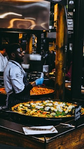 A chef wearing a striped apron and blue gloves is preparing and serving a large pan of colorful paella in a busy market setting. The paella is filled with ingredients like rice, bell peppers, mussels, and boiled eggs. Numerous people are observed in the background, contributing to the lively market atmosphere.