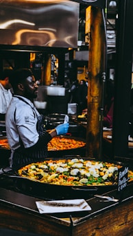 A chef wearing a striped apron and blue gloves is preparing and serving a large pan of colorful paella in a busy market setting. The paella is filled with ingredients like rice, bell peppers, mussels, and boiled eggs. Numerous people are observed in the background, contributing to the lively market atmosphere.