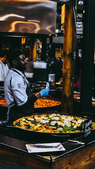 A chef wearing a striped apron and blue gloves is preparing and serving a large pan of colorful paella in a busy market setting. The paella is filled with ingredients like rice, bell peppers, mussels, and boiled eggs. Numerous people are observed in the background, contributing to the lively market atmosphere.