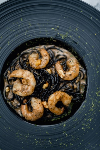 A dish featuring black pasta elegantly arranged on a dark ceramic plate, topped with several well-cooked shrimp. The pasta appears to be garnished with herbs and small nuts, creating a sophisticated and appetizing presentation.