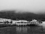 A black and white image features a quaint waterfront scene with a small, elevated restaurant named 'Restaurante D'uportinho' located on a pier. The restaurant is adjacent to a serene body of water with reflections visible on the surface. In the background, a row of traditional houses lines the shore, and dense, fog-covered hills rise behind them, lending a mysterious and tranquil atmosphere to the scene.