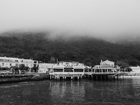 A black and white image features a quaint waterfront scene with a small, elevated restaurant named 'Restaurante D'uportinho' located on a pier. The restaurant is adjacent to a serene body of water with reflections visible on the surface. In the background, a row of traditional houses lines the shore, and dense, fog-covered hills rise behind them, lending a mysterious and tranquil atmosphere to the scene.