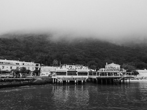 A black and white image features a quaint waterfront scene with a small, elevated restaurant named 'Restaurante D'uportinho' located on a pier. The restaurant is adjacent to a serene body of water with reflections visible on the surface. In the background, a row of traditional houses lines the shore, and dense, fog-covered hills rise behind them, lending a mysterious and tranquil atmosphere to the scene.