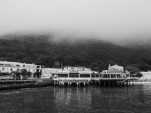 A black and white image features a quaint waterfront scene with a small, elevated restaurant named 'Restaurante D'uportinho' located on a pier. The restaurant is adjacent to a serene body of water with reflections visible on the surface. In the background, a row of traditional houses lines the shore, and dense, fog-covered hills rise behind them, lending a mysterious and tranquil atmosphere to the scene.