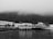 A black and white image features a quaint waterfront scene with a small, elevated restaurant named 'Restaurante D'uportinho' located on a pier. The restaurant is adjacent to a serene body of water with reflections visible on the surface. In the background, a row of traditional houses lines the shore, and dense, fog-covered hills rise behind them, lending a mysterious and tranquil atmosphere to the scene.