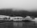 A black and white image features a quaint waterfront scene with a small, elevated restaurant named 'Restaurante D'uportinho' located on a pier. The restaurant is adjacent to a serene body of water with reflections visible on the surface. In the background, a row of traditional houses lines the shore, and dense, fog-covered hills rise behind them, lending a mysterious and tranquil atmosphere to the scene.