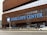 The front entrance of Barclays Center features a large, prominent sign with bold blue lettering against a modern, brown lattice-style facade. The building has a series of glass windows above the signage. A wide road is visible in the foreground, with a few vehicles and pedestrians. A streetlamp is present, adorned with a logo.