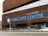 The front entrance of Barclays Center features a large, prominent sign with bold blue lettering against a modern, brown lattice-style facade. The building has a series of glass windows above the signage. A wide road is visible in the foreground, with a few vehicles and pedestrians. A streetlamp is present, adorned with a logo.