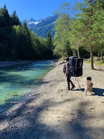A person is walking along a rocky riverbank with a large backpack. Next to the person is a dog on a leash. The river's water is a vibrant blue-green and flows through a lush forested area. In the background, there are towering mountains under a clear blue sky.