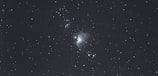 A photograph of a starry night sky with a cloudy nebula near the center. The image showcases numerous small stars scattered throughout the dark expanse.