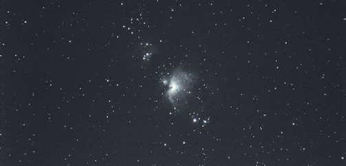 A photograph of a starry night sky with a cloudy nebula near the center. The image showcases numerous small stars scattered throughout the dark expanse.