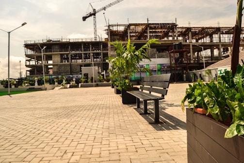 A partially constructed building with scaffolding and a crane towers in the background. In the foreground, there is a paved area lined with benches and decorative plants in planters. Street lights flank the open space, and the sky is partly cloudy.