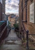 A narrow, cobblestone alleyway descends between two brick buildings. A sign on the right side reads 'Ristorante Collina, Pizzeria, Pasta.' The alley is bordered by railings and leads to a small street lined with residential brick houses. The sky above is partly cloudy, with patches of blue visible.