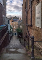 A narrow, cobblestone alleyway descends between two brick buildings. A sign on the right side reads 'Ristorante Collina, Pizzeria, Pasta.' The alley is bordered by railings and leads to a small street lined with residential brick houses. The sky above is partly cloudy, with patches of blue visible.