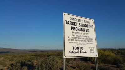A sign stands in a natural landscape under a clear blue sky, indicating that target shooting is prohibited for public safety. It specifies that the area is closed to all target shooting, including air and gas rifles and pistols. The surroundings consist of desert vegetation, including shrubs and cacti, with distant mountains visible on the horizon.