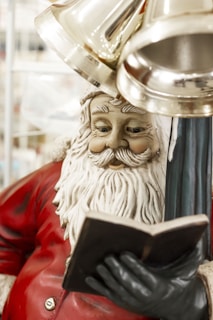 A festive Santa Claus statue holding a book is adorned in a classic red and white outfit, with shiny bells hanging above. The intricate details on Santa’s face and beard, along with the gloved hand holding the book, are well-defined.