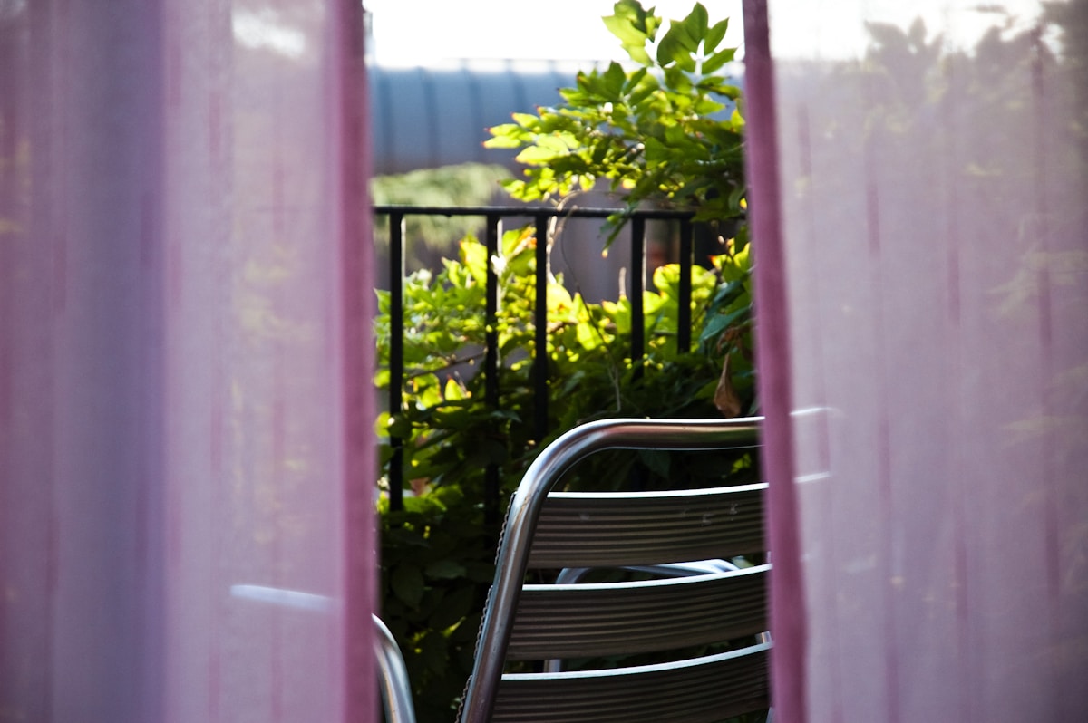 Pink sheer outdoor curtains parted to reveal a small apartment balcony with black metal railing and lush green plants