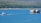A tugboat is seen towing a barge laden with cargo containers across a calm, blue body of water. The background consists of a dense forest on a hillside, and a few small structures can be seen near the shoreline.
