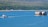 A tugboat is seen towing a barge laden with cargo containers across a calm, blue body of water. The background consists of a dense forest on a hillside, and a few small structures can be seen near the shoreline.