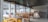 A cozy restaurant interior with wooden tables and colorful chairs, large windows provide a view of a rocky riverbank outside. Minimalist decor includes ceiling lights and a sign that reads 'Wood Fired Oven'.