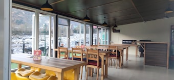 A cozy restaurant interior with wooden tables and colorful chairs, large windows provide a view of a rocky riverbank outside. Minimalist decor includes ceiling lights and a sign that reads 'Wood Fired Oven'.