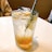 A refreshing beverage in a clear glass, garnished with a sprig of rosemary and lemon wedges. The drink appears to have a gradient with orange at the bottom and clear liquid at the top, filled with ice cubes and bubbles, indicating it's carbonated. The scene suggests a casual dining setting with a blurred background and a tabletop surface.
