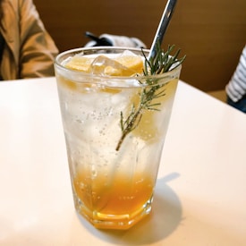 A refreshing beverage in a clear glass, garnished with a sprig of rosemary and lemon wedges. The drink appears to have a gradient with orange at the bottom and clear liquid at the top, filled with ice cubes and bubbles, indicating it's carbonated. The scene suggests a casual dining setting with a blurred background and a tabletop surface.