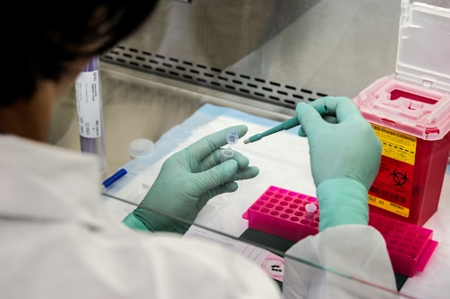 A person wearing green gloves is working in a laboratory setting, using a pipette to transfer liquid into small tubes placed in a pink rack. A red biohazard container is visible on the right side, and various lab equipment and documents are scattered around the workspace.