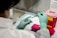 A person wearing green gloves is working in a laboratory setting, using a pipette to transfer liquid into small tubes placed in a pink rack. A red biohazard container is visible on the right side, and various lab equipment and documents are scattered around the workspace.