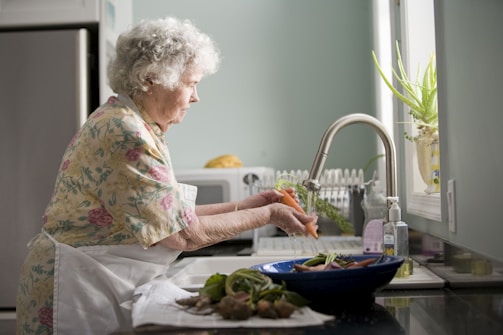An elderly woman with curly white hair wearing a floral-patterned blouse and a white apron washes a carrot under the tap in a kitchen. The kitchen has a soft, pastel green color scheme with a silver refrigerator and a window allowing natural light to enter. An aloe vera plant is situated on the windowsill. On the counter, there are vegetables in a blue bowl and a bottle of liquid soap next to the sink.