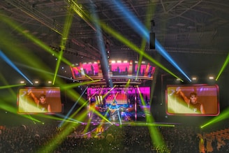 A large-scale concert is taking place in a packed indoor arena. Bright multicolored lights and beams, predominantly green, blue, and pink, illuminate the stage and surrounding area. Two large screens display a close-up of a performer on stage, engaging with the energetic crowd below.