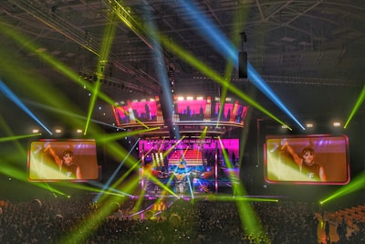 A large-scale concert is taking place in a packed indoor arena. Bright multicolored lights and beams, predominantly green, blue, and pink, illuminate the stage and surrounding area. Two large screens display a close-up of a performer on stage, engaging with the energetic crowd below.