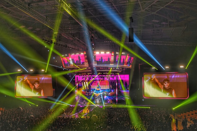 A large-scale concert is taking place in a packed indoor arena. Bright multicolored lights and beams, predominantly green, blue, and pink, illuminate the stage and surrounding area. Two large screens display a close-up of a performer on stage, engaging with the energetic crowd below.