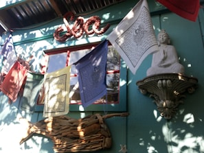 A serene setting with a statue of a meditative figure perched on an ornate shelf against a teal wall, surrounded by colorful prayer flags fluttering in the sunlight. Above them, the word 'Sea' is crafted in a rope-like design. Shadows of leaves playfully dance across the scene, adding a soft layer of texture.