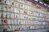 Numerous rows of traditional Japanese sake barrels, each decorated with colorful and intricate designs and Japanese characters, are neatly stacked against a wall. The barrels are wrapped in straw and feature various symbols and images.