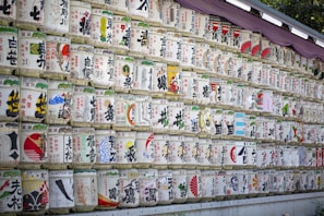 Numerous rows of traditional Japanese sake barrels, each decorated with colorful and intricate designs and Japanese characters, are neatly stacked against a wall. The barrels are wrapped in straw and feature various symbols and images.