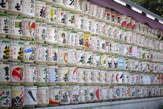Numerous rows of traditional Japanese sake barrels, each decorated with colorful and intricate designs and Japanese characters, are neatly stacked against a wall. The barrels are wrapped in straw and feature various symbols and images.