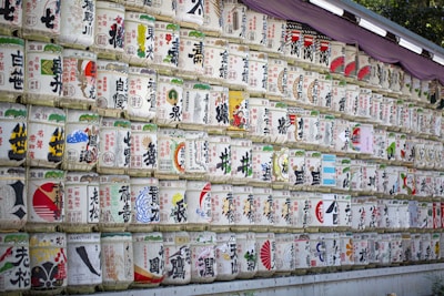 Numerous rows of traditional Japanese sake barrels, each decorated with colorful and intricate designs and Japanese characters, are neatly stacked against a wall. The barrels are wrapped in straw and feature various symbols and images.