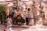 An ornate and vibrant Balinese ceremonial scene with elaborately dressed performers, possibly part of a traditional dance or ritual, set in a temple courtyard. Intricately designed masks, costumes with gold and red accents, and traditional parasols enhance the cultural atmosphere. The surroundings include stone carvings and tropical foliage.
