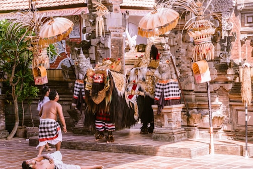 An ornate and vibrant Balinese ceremonial scene with elaborately dressed performers, possibly part of a traditional dance or ritual, set in a temple courtyard. Intricately designed masks, costumes with gold and red accents, and traditional parasols enhance the cultural atmosphere. The surroundings include stone carvings and tropical foliage.