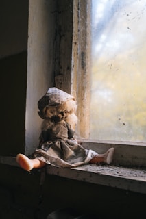 A worn doll with a weathered dress and headscarf sits on a dusty windowsill. The scene is illuminated by soft, natural light filtering through the dirty window, creating a nostalgic and slightly eerie atmosphere.