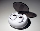 A pair of wireless earbuds resting in an open charging case. The case is white, oval-shaped with a smooth, glossy finish, and a transparent lid. The earbuds have circular touch controls highlighted with a red rim.