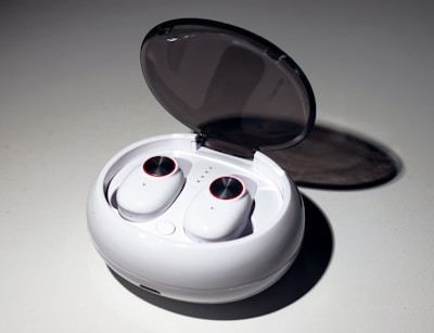 A pair of wireless earbuds resting in an open charging case. The case is white, oval-shaped with a smooth, glossy finish, and a transparent lid. The earbuds have circular touch controls highlighted with a red rim.