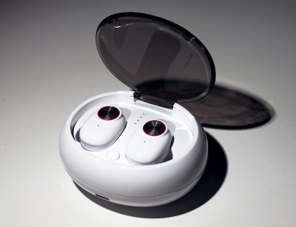 A pair of wireless earbuds resting in an open charging case. The case is white, oval-shaped with a smooth, glossy finish, and a transparent lid. The earbuds have circular touch controls highlighted with a red rim.