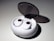 A pair of wireless earbuds resting in an open charging case. The case is white, oval-shaped with a smooth, glossy finish, and a transparent lid. The earbuds have circular touch controls highlighted with a red rim.