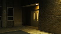 A dimly lit exterior corner of a building featuring a closed double door with narrow windows. The area is surrounded by brick walls, and the ground appears to be paved. A sign indicating handicapped parking is visible to the left near a window.