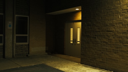 A dimly lit exterior corner of a building featuring a closed double door with narrow windows. The area is surrounded by brick walls, and the ground appears to be paved. A sign indicating handicapped parking is visible to the left near a window.
